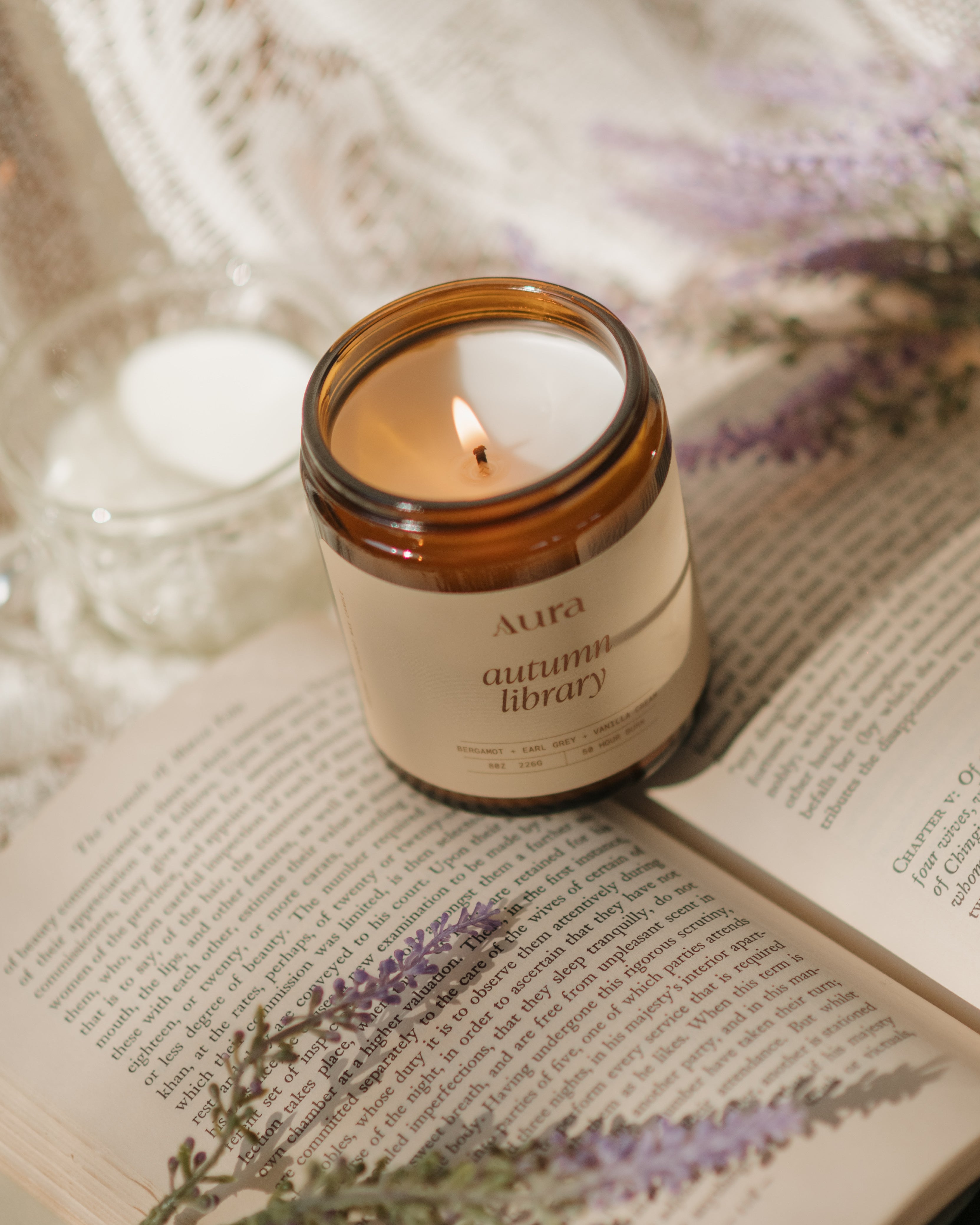 An amber glass jar candle labeled 'Aura autumn library' sits on an open book with purple flowers, styled with a white lace cloth and a small lit candle in the background.