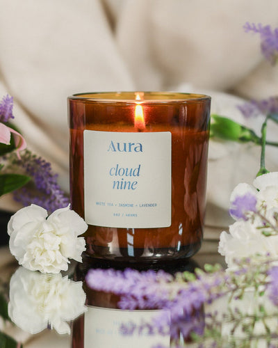 A lit amber glass jar candle with a white label reading 'Aura cloud nine' sits on a reflective surface surrounded by soft pink, white, and purple flowers against a warm cream fabric background.